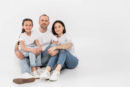 glückliche Familie in weißen T-Shirts und blauen Jeans, in die Kamera blickend und gemeinsam auf grauem Hintergrund sitzend, Internationaler Kinderschutztag, Eltern und Tochter 
