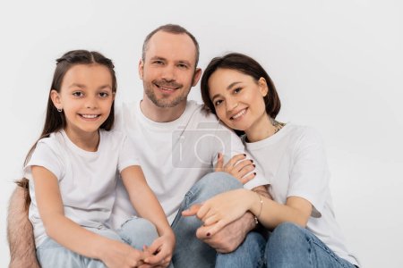portrait of happy family in white t-shirts and blue denim jeans looking at camera and sitting together on grey background, International child protection day, parents and daughter 