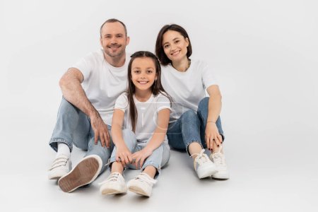 Stilvolle Familie in weißen T-Shirts und blauen Jeans, die in die Kamera blickt und vor grauem Hintergrund zusammensitzt, Internationaler Kinderschutztag, glückliche Eltern und Tochter 