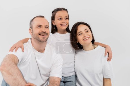 stylische Familie in weißen T-Shirts, die wegguckt, während sie gemeinsam auf grauem Hintergrund posiert, Internationaler Kinderschutztag, Frühchen-Tochter umarmt glückliche Eltern 