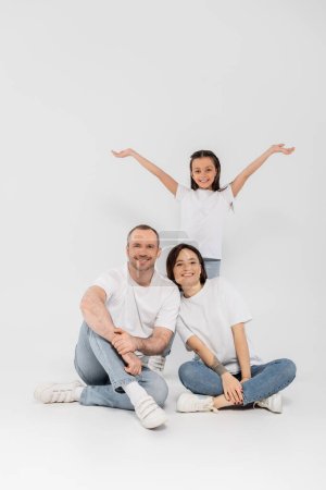 Frühchen-Tochter steht mit ausgestreckten Händen neben glücklichen Eltern in weißen T-Shirts und blauen Jeans und blickt in die Kamera und posiert gemeinsam auf grauem Hintergrund, Kinderschutztag 