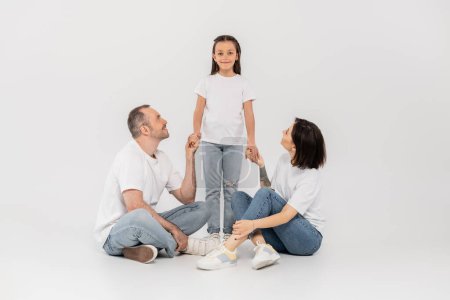 Glückliche Eltern in weißen T-Shirts und blauen Jeans sitzend und Händchen haltend, die lächelnde Frühchen-Tochter vor grauem Hintergrund in die Kamera blickend, Tag des Kinderschutzes, 1. Juni
