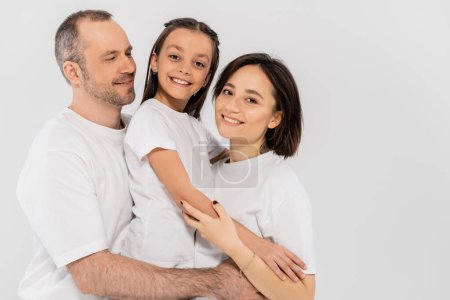 portrait of happy family in white t-shirts looking at camera and hugging each other on grey background, International child protection day, parents and daughter 