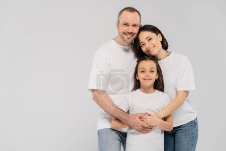 happy parents hugging cheerful daughter while standing together in blue denim jeans and white t-shirts and looking at camera on grey background, Happy children's day, June 1