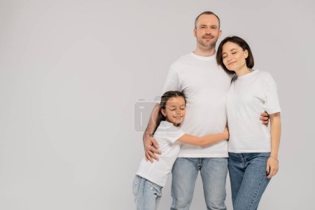 Photo for Happy preteen girl hugging father near mother with tattoo while standing together in blue denim jeans and white t-shirts and looking at camera on grey background, Happy children's day - Royalty Free Image