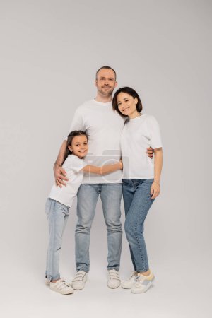 joyous preteen girl hugging father near mother with tattoo while standing together in blue denim jeans and white t-shirts and looking at camera on grey background, Happy children's day, 