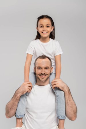 happy preteen daughter with brunette hair sitting on shoulders of unshaved and cheerful father with bristle and looking at camera on grey background, Child protection day 