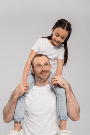 glückliche Tochter mit langen brünetten Haaren sitzt auf den Schultern des unrasierten und fröhlichen Vaters mit Borsten auf grauem Hintergrund, Kinderschutztag, Vater und Tochter 