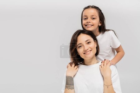 happy preteen daughter with long brunette hair holding hands of cheerful mother with tattoo and looking at camera on grey background, Child protection day, mother and daughter 