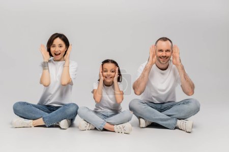 amazed father and tattooed mother with short hair and excited preteen daughter sitting with crossed legs in white t-shirts and blue denim jeans on grey background, Happy children's day 