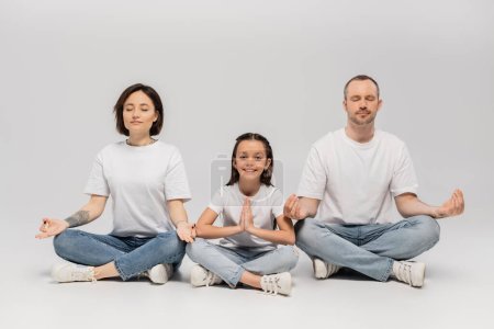 Vater und tätowierte Mutter mit kurzen Haaren meditieren mit fröhlicher Tochter, während sie mit überkreuzten Beinen in weißen T-Shirts und blauen Jeans auf grauem Hintergrund sitzen 
