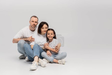 freudiger Vater und Mutter mit kurzen Haaren, die fröhliche Tochter umarmt, während sie mit gekreuzten Beinen in weißen T-Shirts und blauen Jeans auf grauem Hintergrund sitzt, Froher Kindertag 