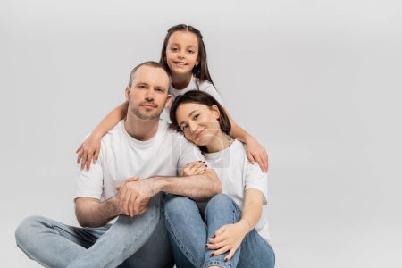joyous preteen girl hugging cheerful parents in white t-shirts and blue denim jeans while bonding together and looking at camera on grey background, Happy children's day 