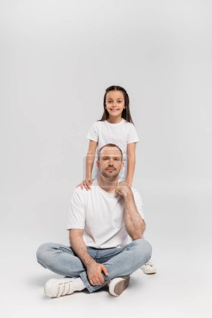 glückliches Mädchen mit brünetten Haaren umarmt fröhlichen Vater mit Borsten, während sie in weißen T-Shirts und blauen Jeans posiert und in die Kamera auf grauem Hintergrund schaut, Happy Vatertag
