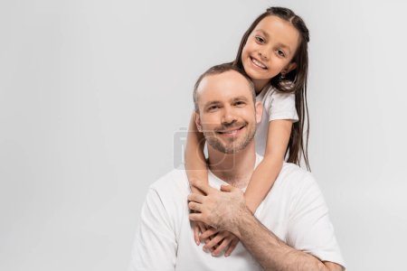 portrait de fille heureuse avec de longs cheveux bruns étreignant père joyeux avec soies tout en posant dans des t-shirts blancs et en regardant la caméra sur fond gris, Bonne fête des pères