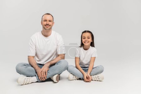 happy preteen girl with long brunette hair and cheerful dad with bristle sitting with crossed legs while posing in white t-shirts and blue denim jeans on grey background, Happy Father`s Day
