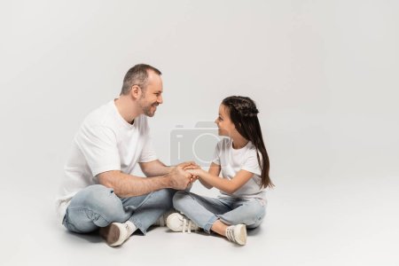 glückliches Mädchen mit langen brünetten Haaren, das mit fröhlichem Papa Händchen hält, während es mit überkreuzten Beinen in weißen T-Shirts und blauen Jeans auf grauem Hintergrund sitzt, Happy Father 's Day