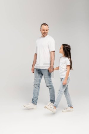 Ein fröhlicher Vater mit Borsten und eine frühpubertäre Tochter in weißen T-Shirts und blauen Jeans, die sich an den Händen halten und gemeinsam auf grauem Grund spazieren gehen.