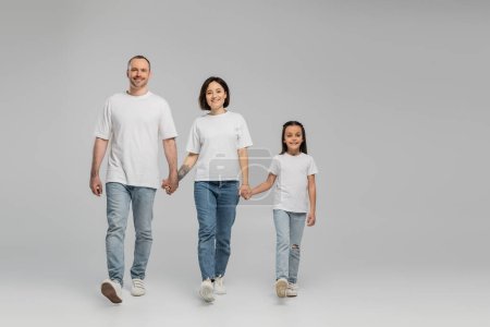 longitud completa de padres felices e hija en camisetas blancas y vaqueros azules tomados de la mano y caminando juntos sobre fondo gris, Día Internacional de la Protección del Niño en junio 