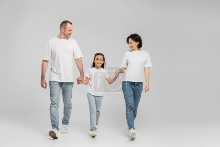 full length of happy parents and kid in white t-shirts and blue denim jeans holding hands and walking together on grey background, International child protection day in June 