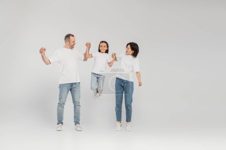 full length of happy parents in white t-shirts and blue denim jeans holding hands and lifting preteen daughter while standing together on grey background, International child protection day in June 
