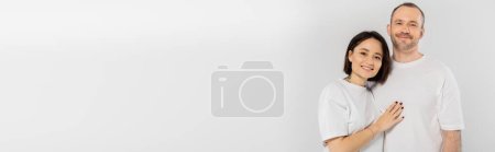 Foto de Mujer tatuada con pelo corto morena abrazando alegre marido con cerdas mientras están de pie juntos en camisetas blancas y mirando a la cámara aislada sobre fondo gris, feliz pareja, pancarta - Imagen libre de derechos