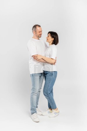 volle Länge der lächelnden Frau mit kurzen brünetten Haaren umarmt Ehemann mit Borsten, während sie zusammen in weißen T-Shirts und Jeans stehen und einander auf grauem Hintergrund anschauen, glückliches Paar 