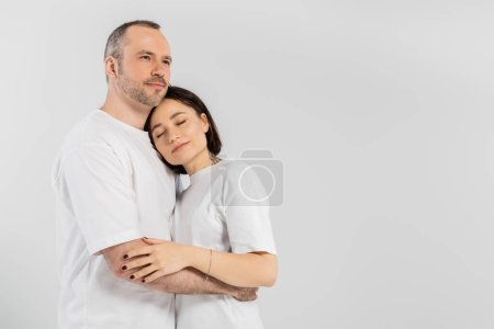 tattooed woman with short brunette hair and closed eyes leaning on chest of husband with bristle while standing together in white t-shirts isolated on grey background, happy couple 