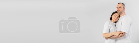 Photo for Tattooed woman with short brunette hair leaning on chest of husband while standing together in white t-shirts isolated on grey background, happy couple, banner - Royalty Free Image