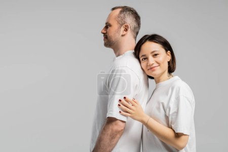 tätowierte Frau mit kurzen brünetten Haaren, die sich auf den Rücken ihres Mannes lehnt, während sie in weißen T-Shirts zusammensteht und vereinzelt in die Kamera auf grauem Hintergrund blickt, glückliches Paar 