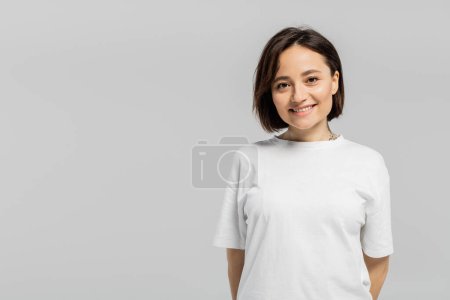 happy and tattooed woman with short hair and natural makeup standing in white t-shirt and smiling while looking at camera isolated on grey background with copy space 
