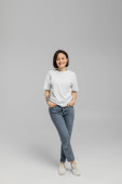 full length of happy and tattooed woman with short hair and natural makeup standing in white t-shirt and posing with hands in pockets of blue denim jeans while looking at camera on grey background  Poster #656741316