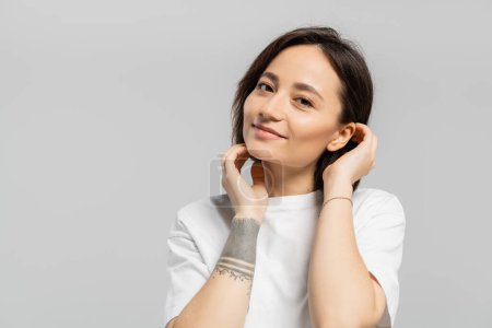 mujer atractiva y tatuada tocando su pelo corto y de pie en camiseta blanca, sonriendo mientras mira a la cámara aislada sobre fondo gris con espacio para copiar 