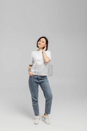 full length of appealing and tattooed woman with short hair and natural makeup standing in white t-shirt and posing with hand in pocket of blue denim jeans while looking at camera on grey background 