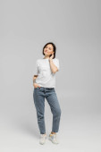 full length of appealing and tattooed woman with short hair and natural makeup standing in white t-shirt and posing with hand in pocket of blue denim jeans while looking at camera on grey background  Tank Top #656741384