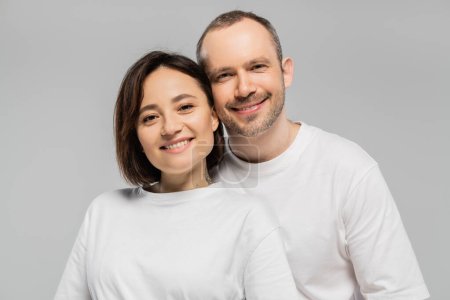 cheerful husband and tattooed wife with short brunette hair standing together in white t-shirts and looking at camera isolated on grey background in studio, happy couple 