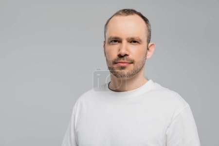 unshaved man with bristle standing in white t-shirt and looking at camera while posing isolated on grey background in studio, copy space, confidence and masculinity 