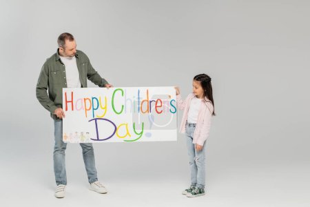 Frühchen Mädchen mit langen brünetten Haaren und Vater und legerer Kleidung halten Plakat mit Happy Children 's Day Worten und stehen zusammen auf grauem Hintergrund, Kinderschutztag, Studio 