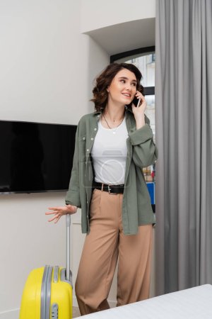 positive woman with brunette hair, in casual clothes, shirt and pants, talking on mobile phone while standing near yellow suitcase, window, grey curtains and lcd tv with blank screen in hotel room