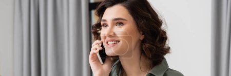phone call, young and excited woman with brunette hair talking on mobile phone near grey curtains in contemporary and cozy hotel suite, leisure and travel, banner