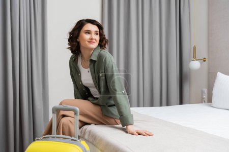young woman with brunette hair, wearing casual shirt and pants, sitting on bed near yellow suitcase, looking away and smiling in comfortable hotel room, happy traveler, weekend break
