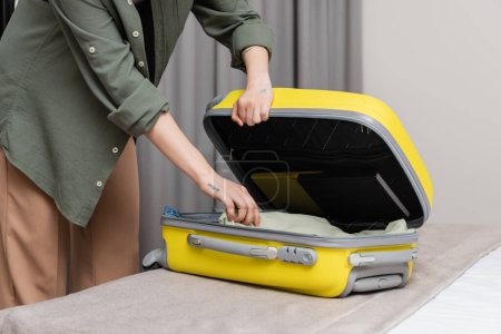 Photo for Partial view of young tattooed woman in casual clothes unpacking yellow suitcase with travel necessities on comfortable bed in modern hotel room near grey curtains on blurred background - Royalty Free Image