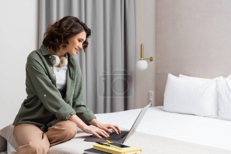 digitaler Nomadentum, lächelnde tätowierte Frau mit drahtlosen Kopfhörern, die auf dem Bett sitzt und in der Nähe von Notebooks, Stift-Smartphones, weißen Kissen, Wandleuchte und grauen Vorhängen im Hotelzimmer arbeitet
