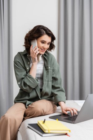 cheerful tattooed woman with wireless headphones talking on mobile phone and working on laptop while sitting near grey curtains, notebooks and pen on bed in comfortable hotel room, phone call 
