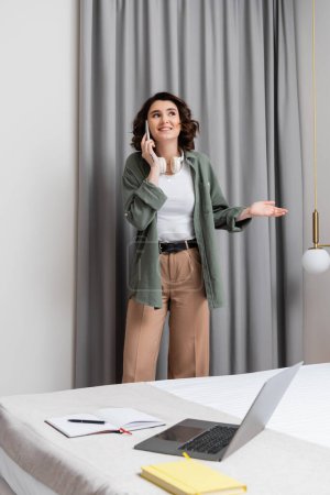 cheerful tattooed woman with wavy brunette hair and headphones talking on smartphone near grey curtains, wall sconce, laptop, notebooks and pen on bed in modern hotel room, work-life balance