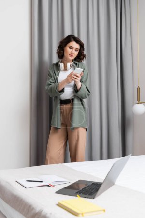 appealing woman with wireless headphones standing near grey curtains, wall sconce, bed with laptop, notebooks and pen, browsing internet on mobile phone in cozy atmosphere of hotel suite