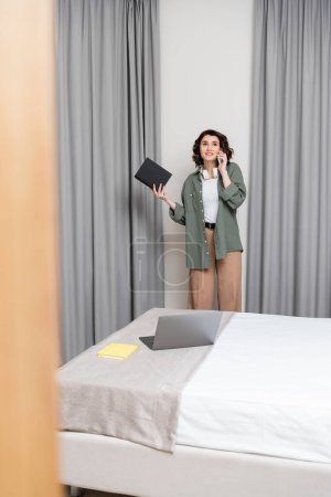 freelance lifestyle, happy young woman in casual clothes with wireless headphones and notebook talking on mobile phone while standing near grey curtains and bed with notepad and laptop in hotel room 