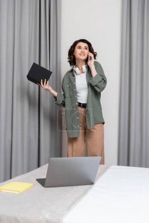 pleased woman with wavy brunette hair and wireless headphones holding notebook, looking away and talking on smartphone near grey curtains and laptop with notepad on comfortable bed in hotel room