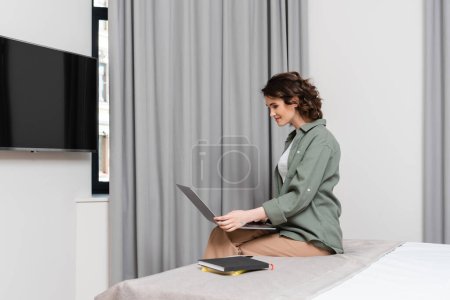 Téléchargez les photos : Femme joyeuse en vêtements décontractés, avec des cheveux bruns ondulés assis sur le lit et regardant ordinateur portable près de lcd tv, rideaux gris et cahiers dans une suite d'hôtel confortable et moderne, travail et Voyage - en image libre de droit