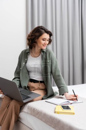 zufriedene junge Frau mit welligem brünetten Haar sitzt mit Laptop im Bett und schreibt in Notizbuch neben Smartphone mit leerem Bildschirm, Notizblock und grauen Vorhängen in komfortablem Hotelzimmer